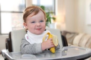 a baby eating a banana at his feeding table wearing a Bibby bib