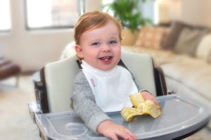 a smiling baby holding a banana at his feeding table wearing a Bibby bib