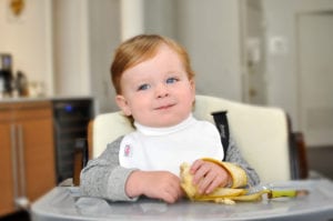 a baby eating a banana at his feeding table wearing a Bibby bib