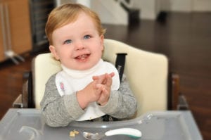 a toddler sitting at his feeding table smiling and wearing a Bibby bib